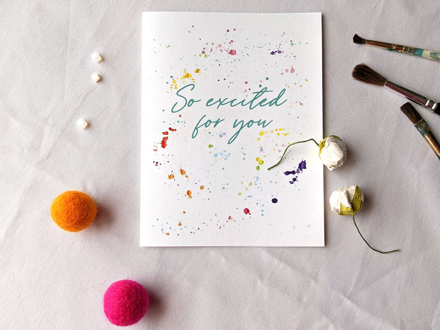 Greeting card laying on a white background surrounded by white beads, colorful felt balls, paintbrushes and two faux flower buds.