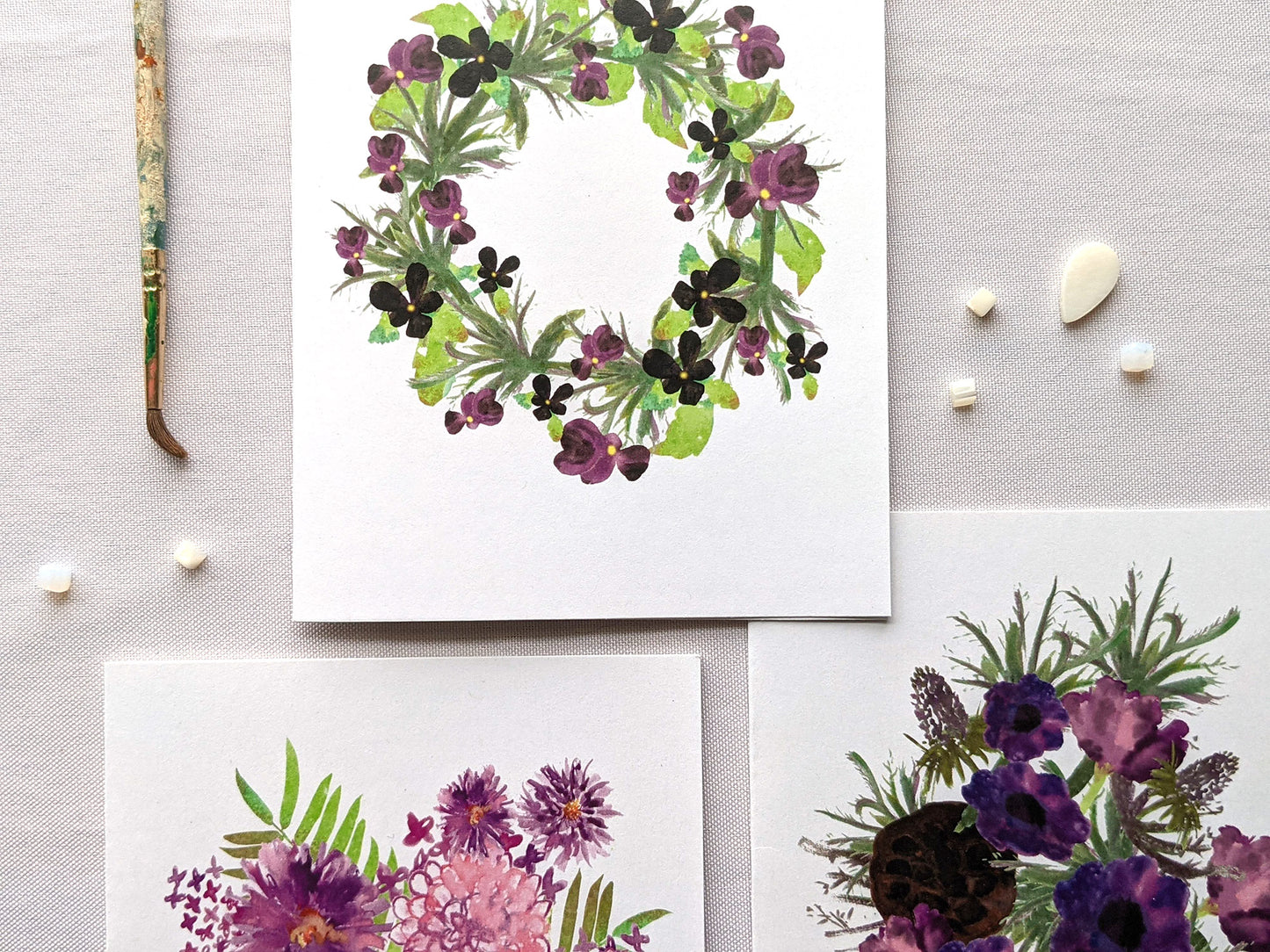 Overlapping greeting cards on a white background next to beads and paintbrush.