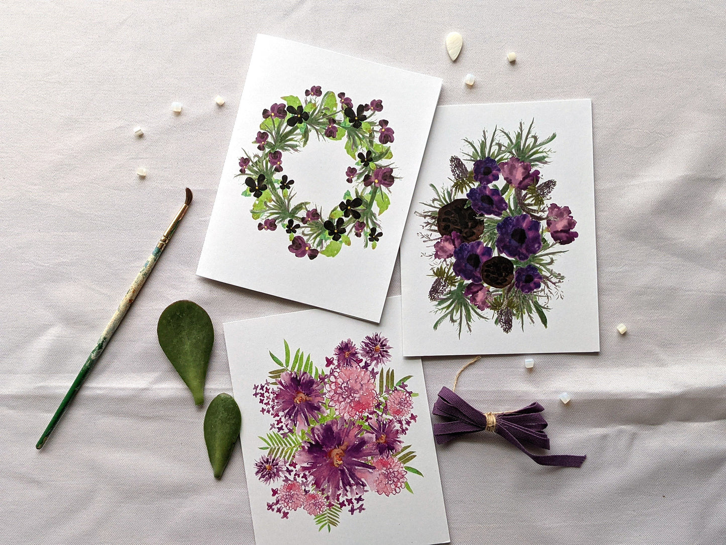 Overlapping greeting cards on a white background next to beads, paintbrush, succulent leaves, and colorful cotton ribbon.