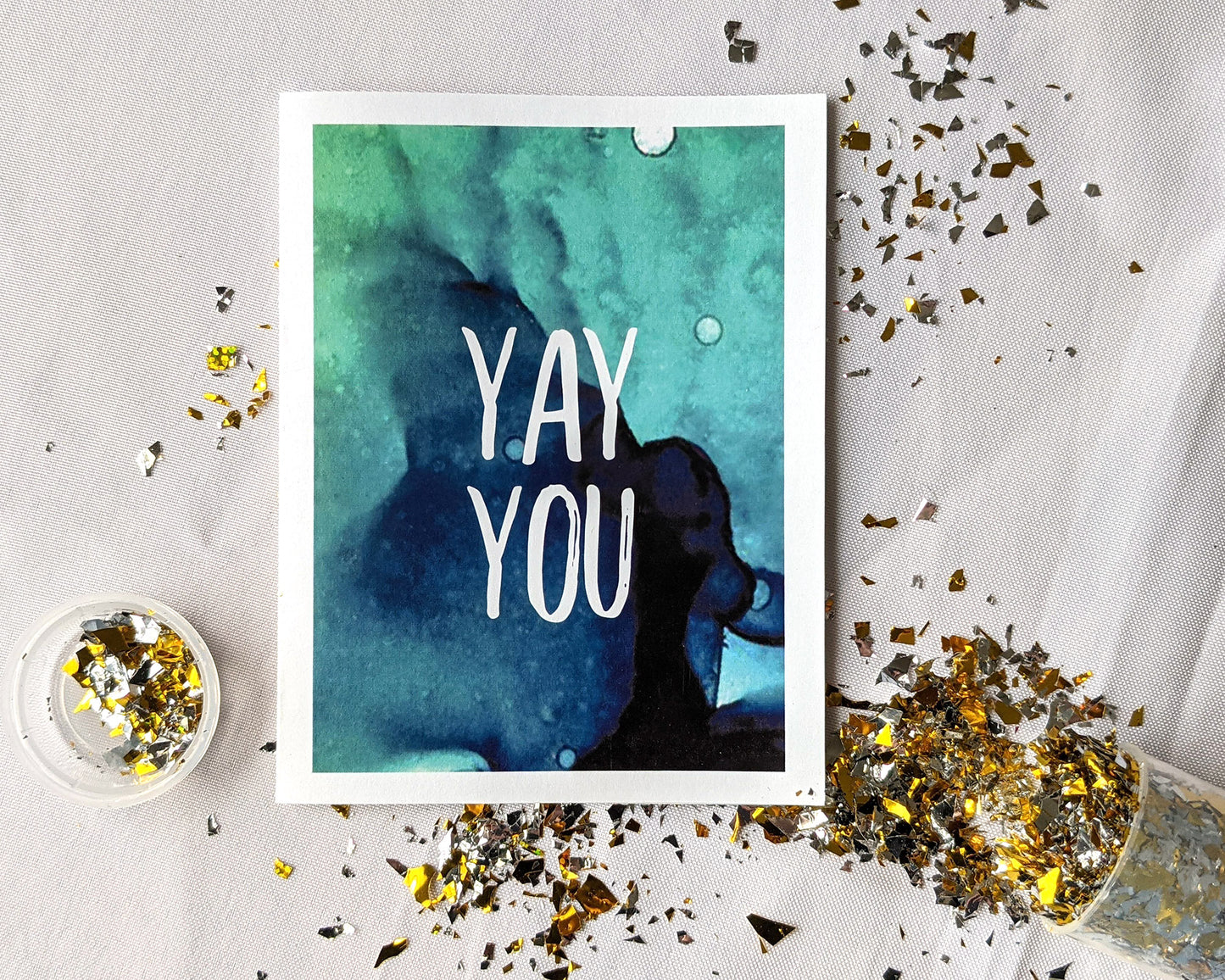 Greeting card laying on a white background surrounded by spilled gold confetti and its cannister.