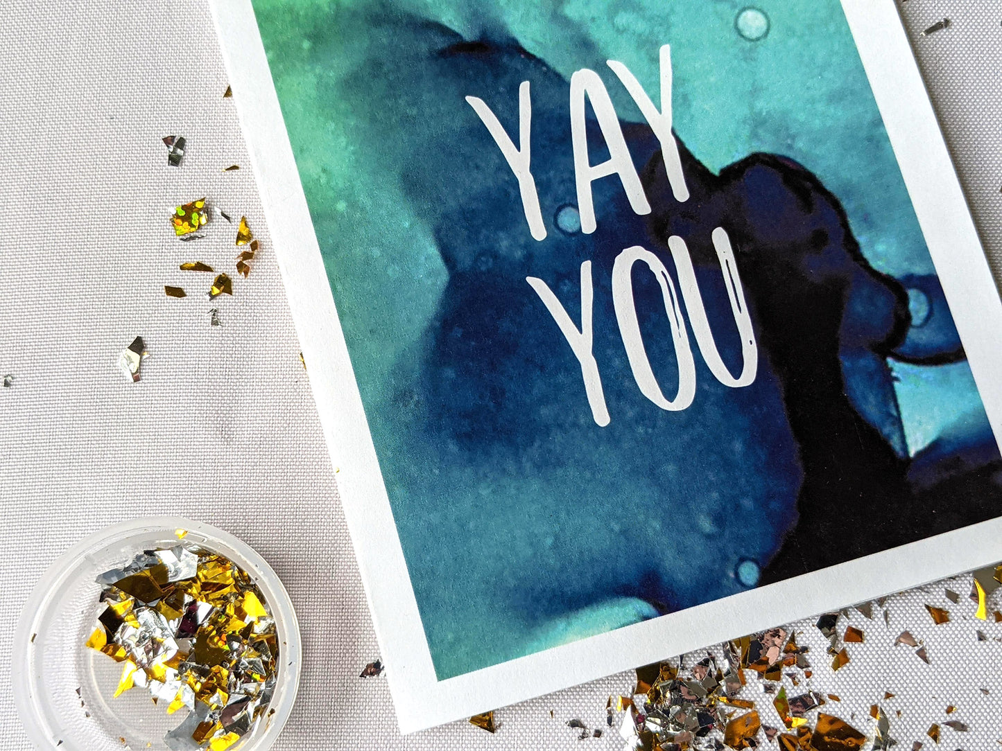 Closeup of a greeting card laying on a white background surrounded by spilled gold confetti and its cannister.