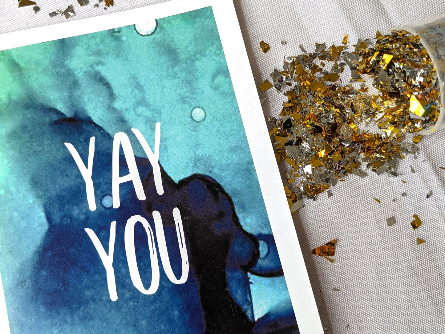 Closeup of a greeting card laying on a white background surrounded by spilled gold confetti and its cannister.