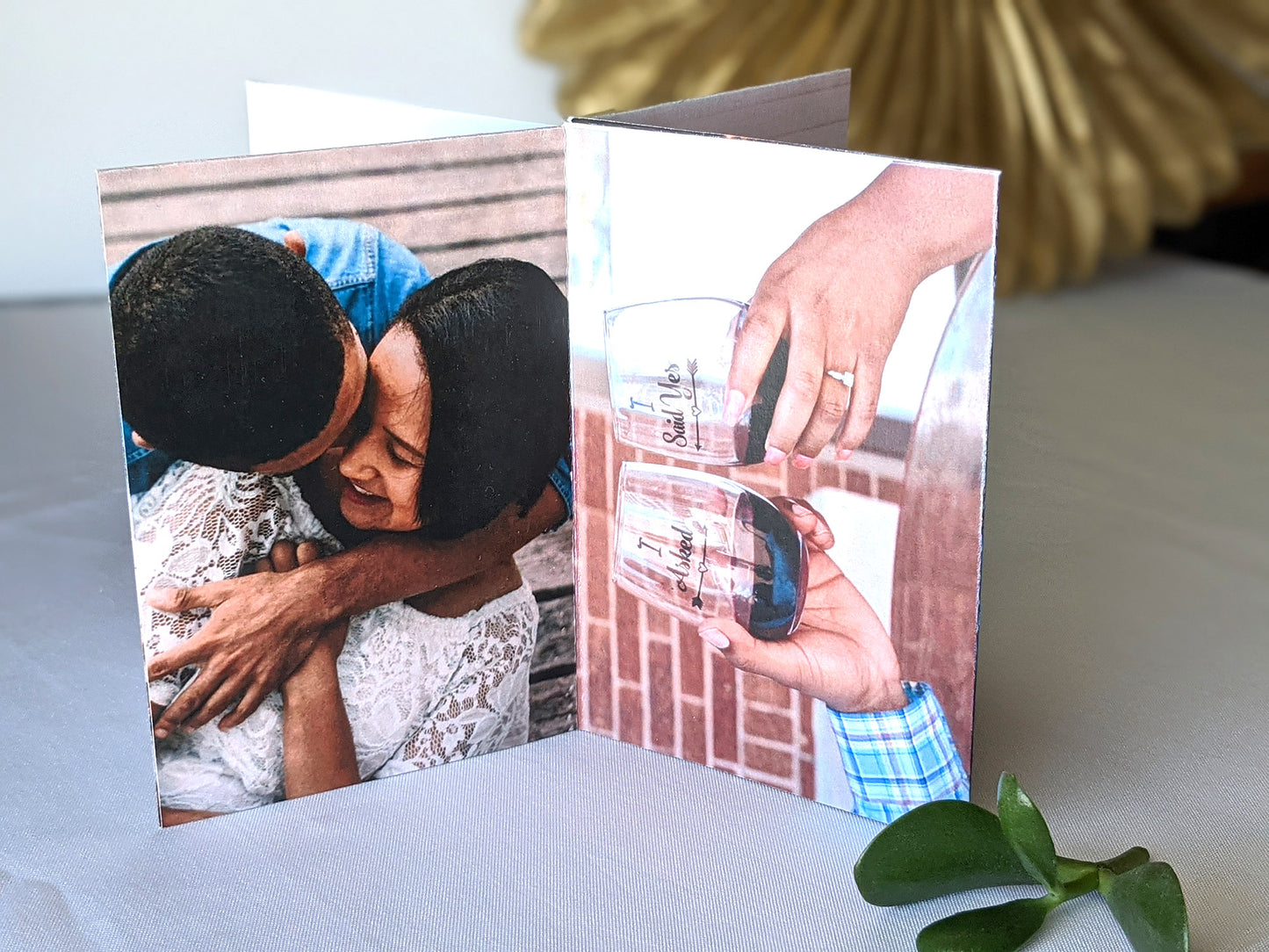 Centerpiece showing a couple smiling at each other with their heads close and arms wrapped and clinking personalized drinking glasses with their hands showcasing a ring sitting on a white tablecloth with a gold paper medallion and green plant leaves off to the side.
