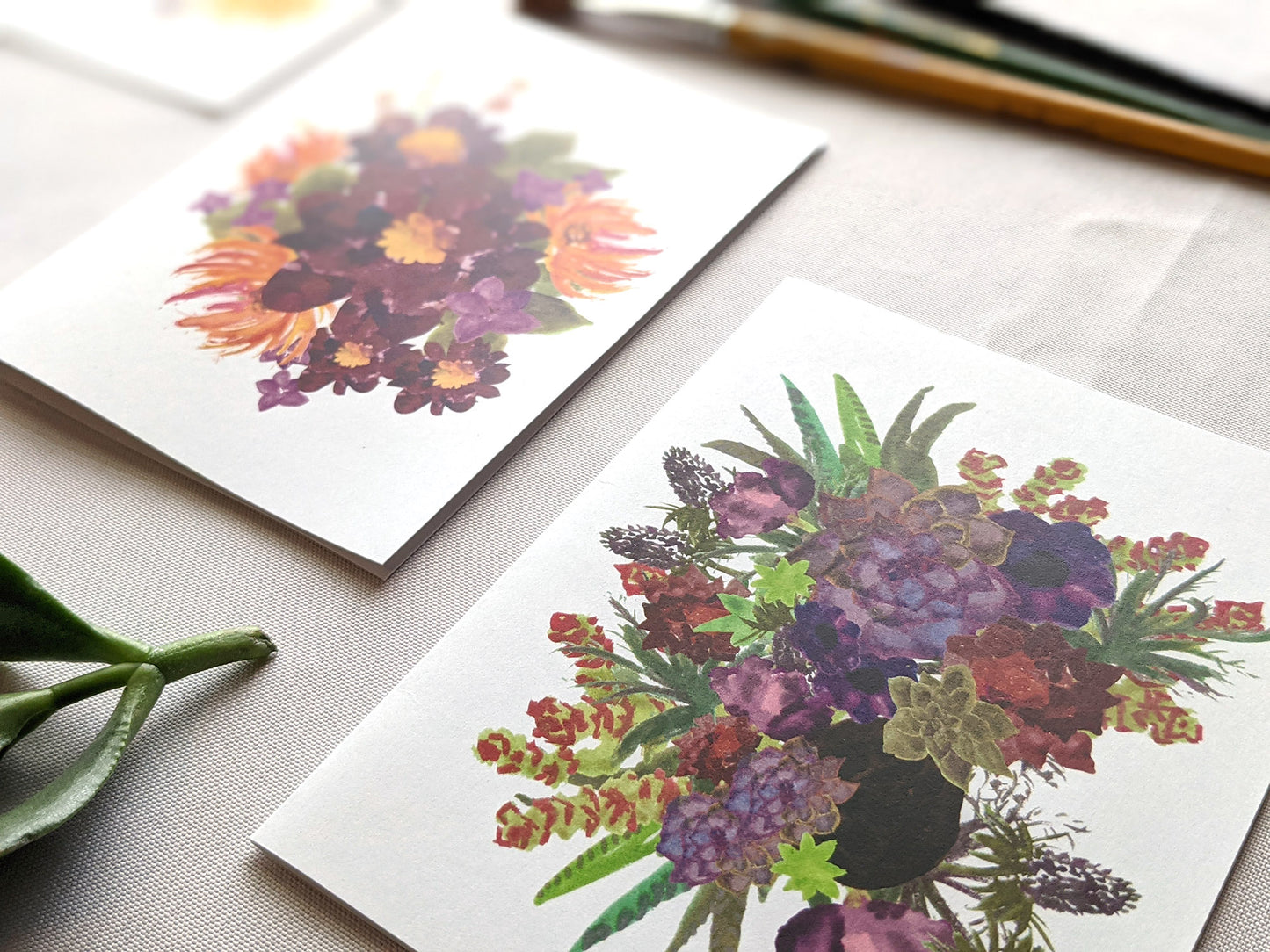 Angled view of greeting cards laying on a white surface with succulent leaves and paint brushes.