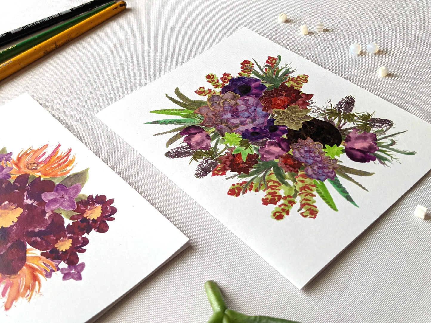 Angled view of greeting cards laying on a white surface with succulent leaves, paint brushes and beads.