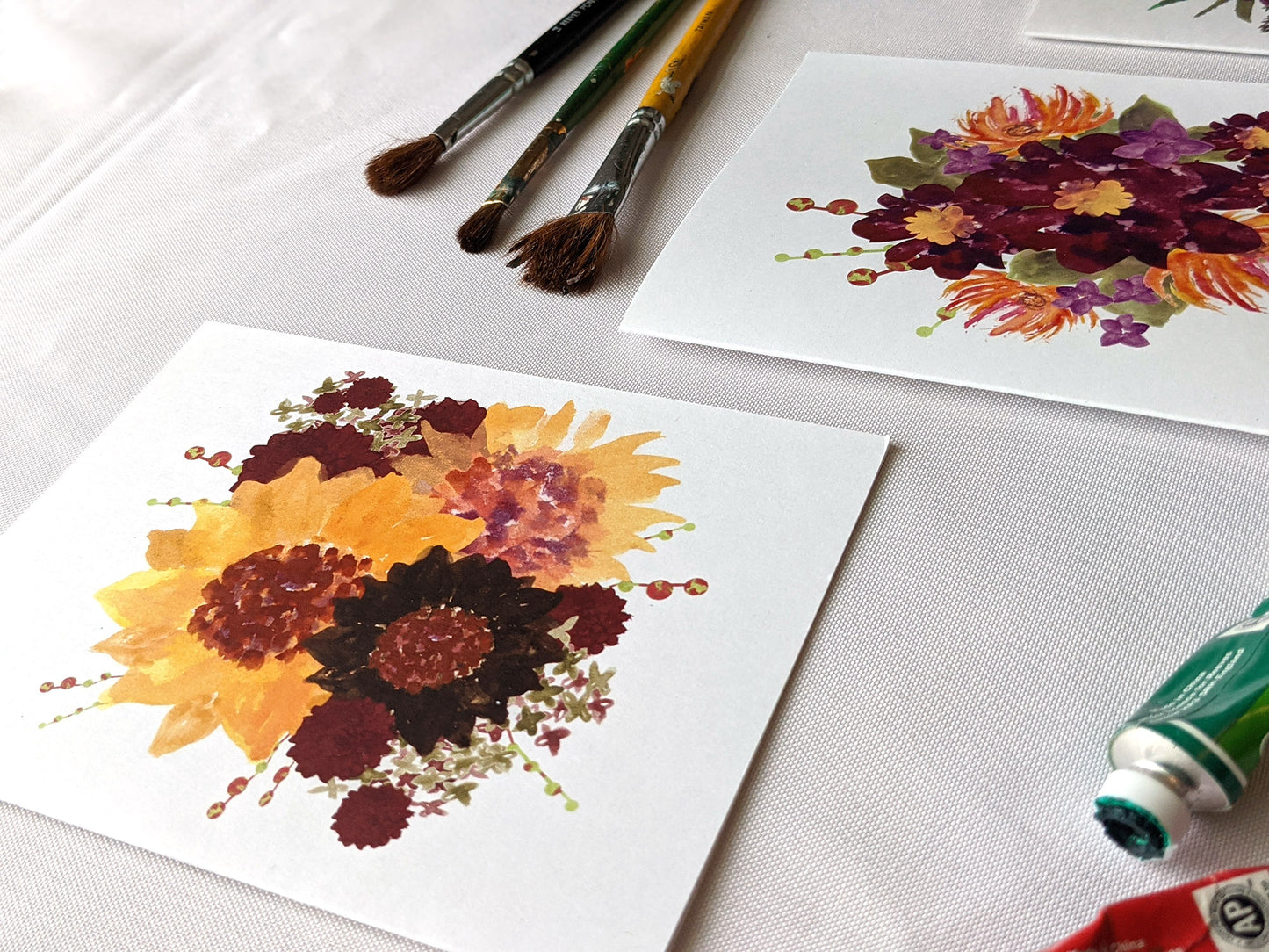 Angled view of greeting cards on a white surface with paintbrushes and paint tubes.