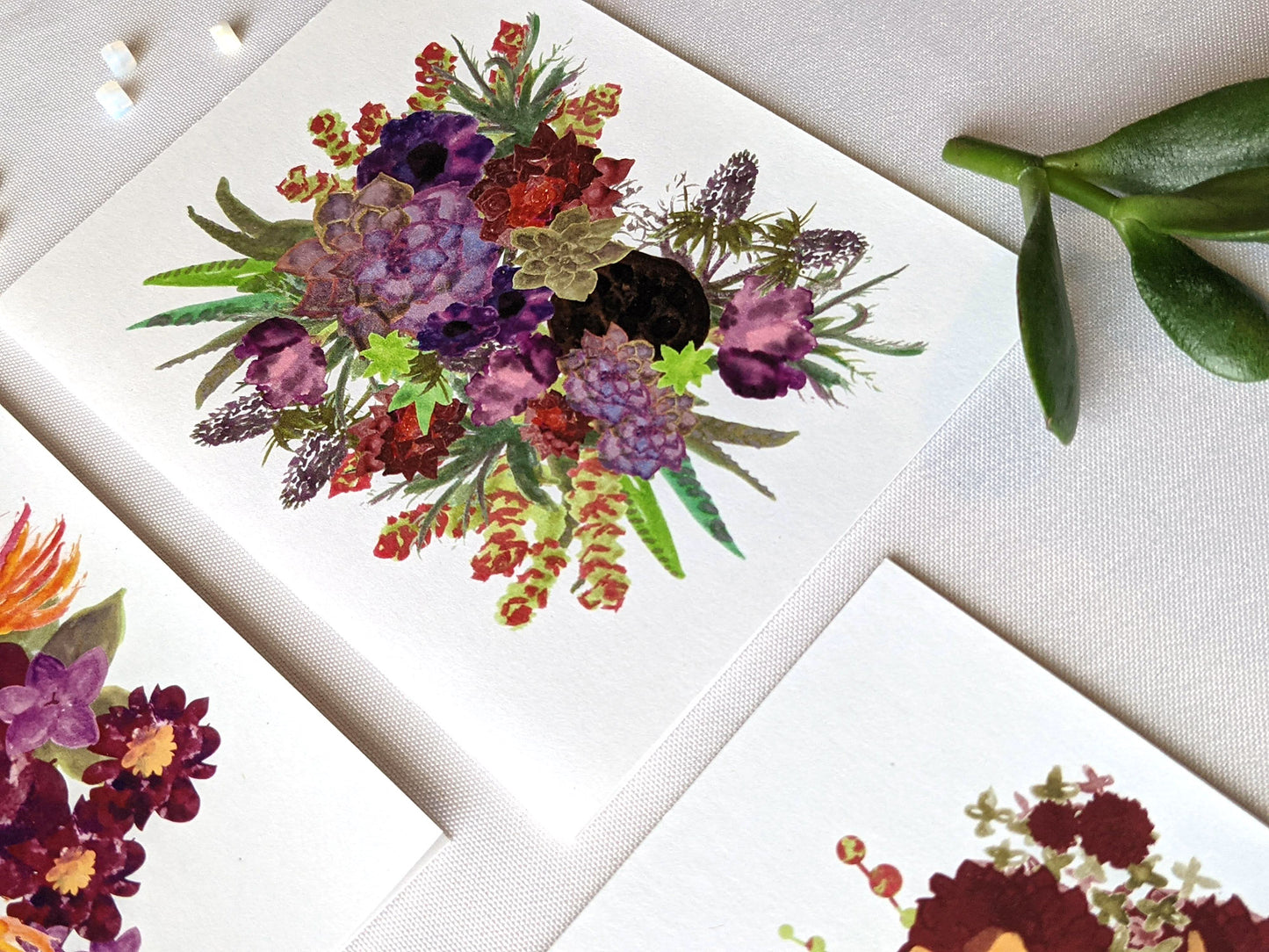 Angled view of greeting cards laying on a white surface with succulent leaves and beads.