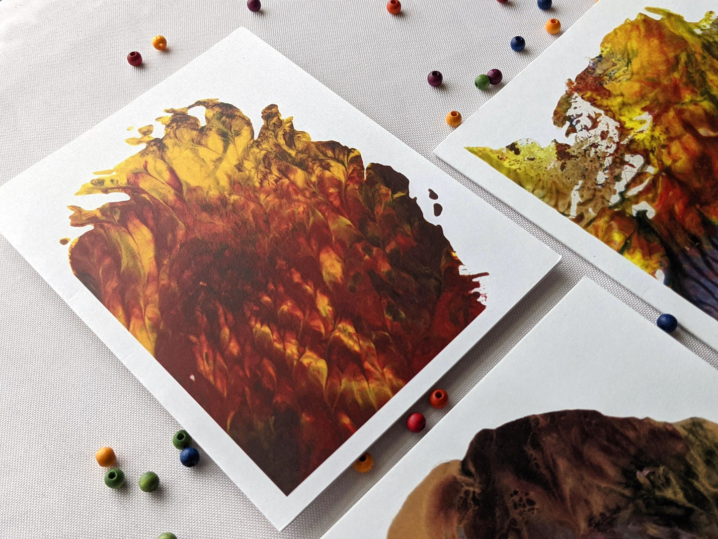 Three greeting cards with abstract paintings laying on a white cloth background surrounded by small colorful wooden beads.