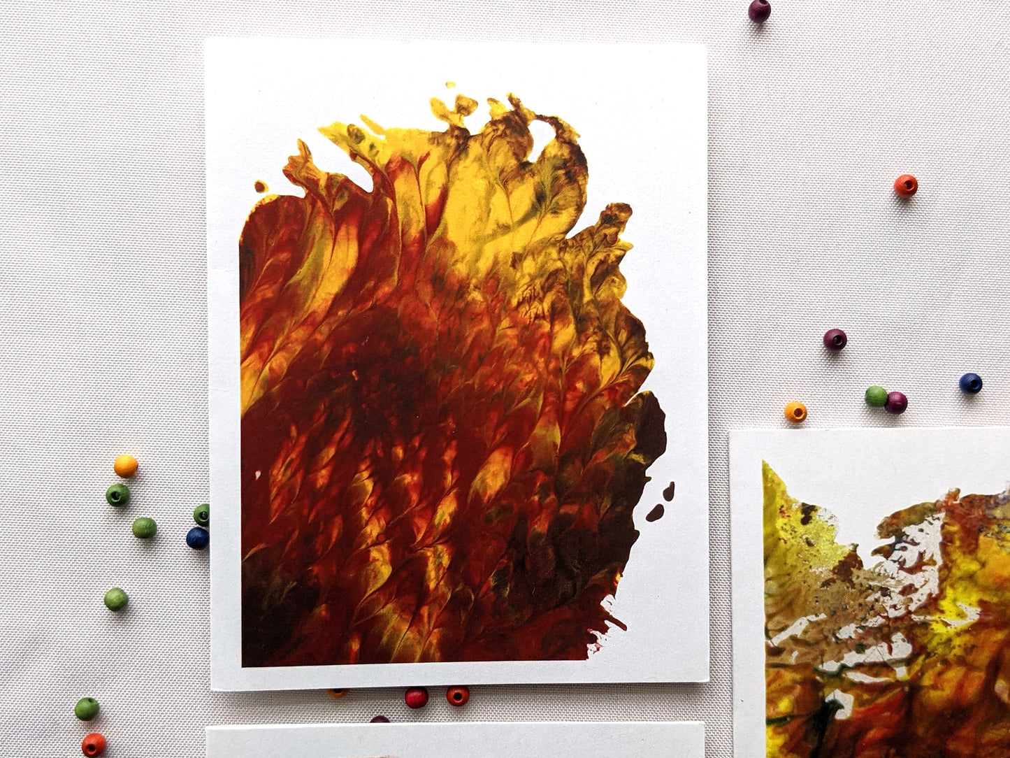 Greeting card showing an abstract painting in reds, yellows, and browns created from squishing acrylic paint between pieces of cardboard. It sits on white background with colorful wooden beads and the edges of two other greeting cards.