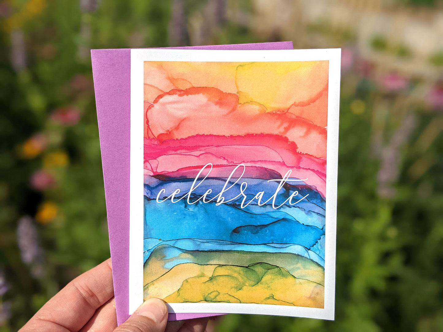 Hand holding a greeting card outdoors with pink, purple, and yellow flowers blurred in the background.