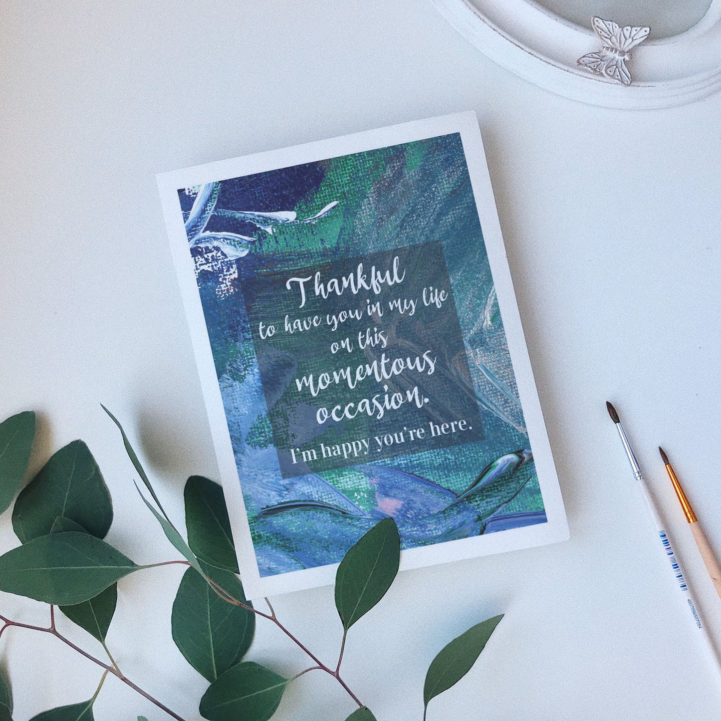 Greeting card laying on a white background near green leaves, paintbrushes and a ceramic dish.