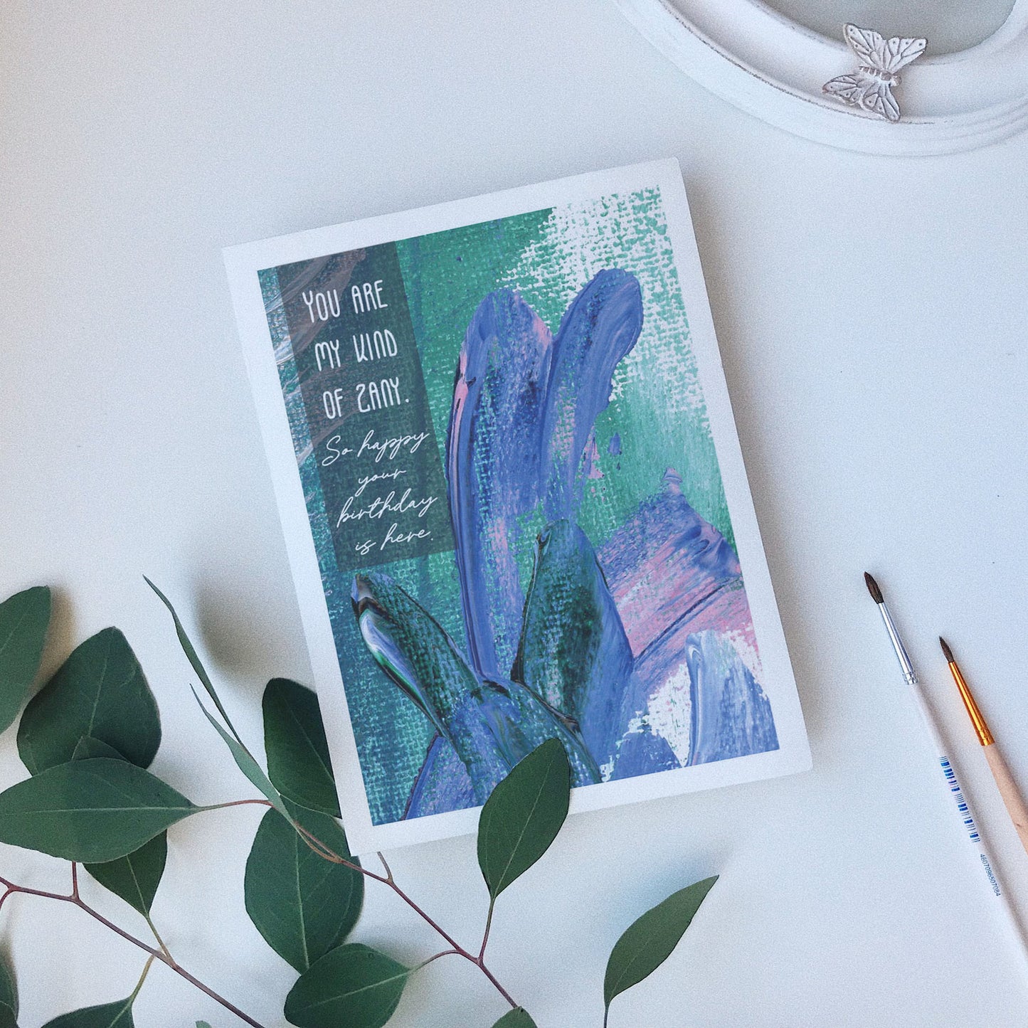 Greeting card laying on a white background near green leaves, paintbrushes and a ceramic dish.