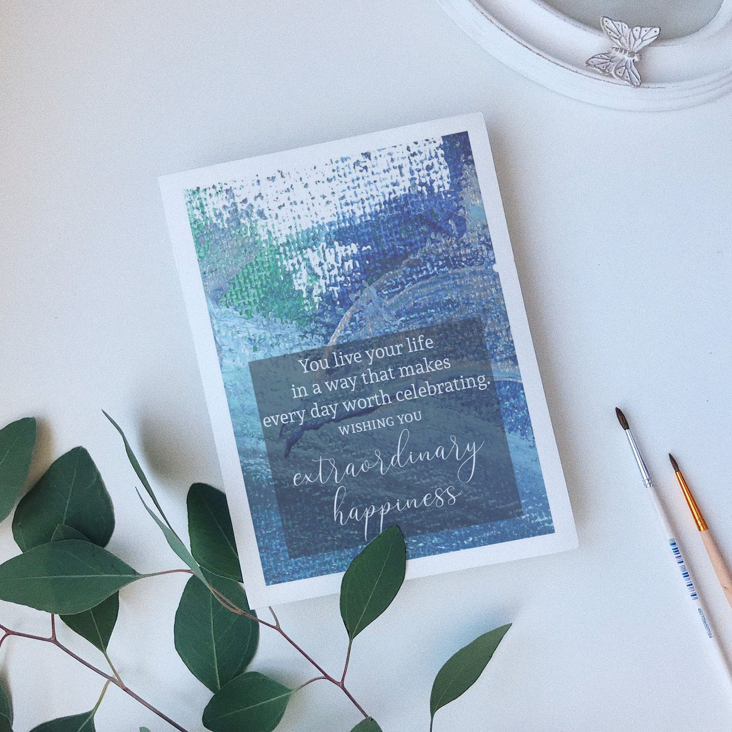 Greeting card laying on a white background near green leaves, paintbrushes and a ceramic dish.