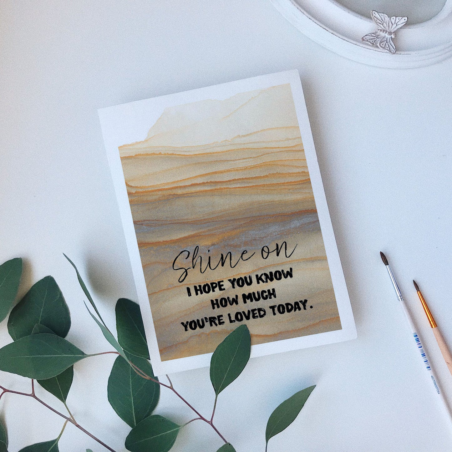 Greeting card laying on a white background near green leaves, paintbrushes and a ceramic dish.