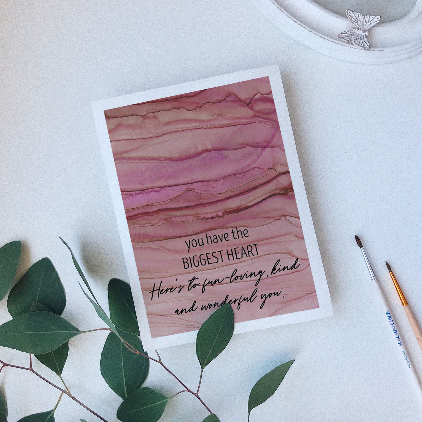 Greeting card laying on a white background near green leaves, paintbrushes and a ceramic dish.