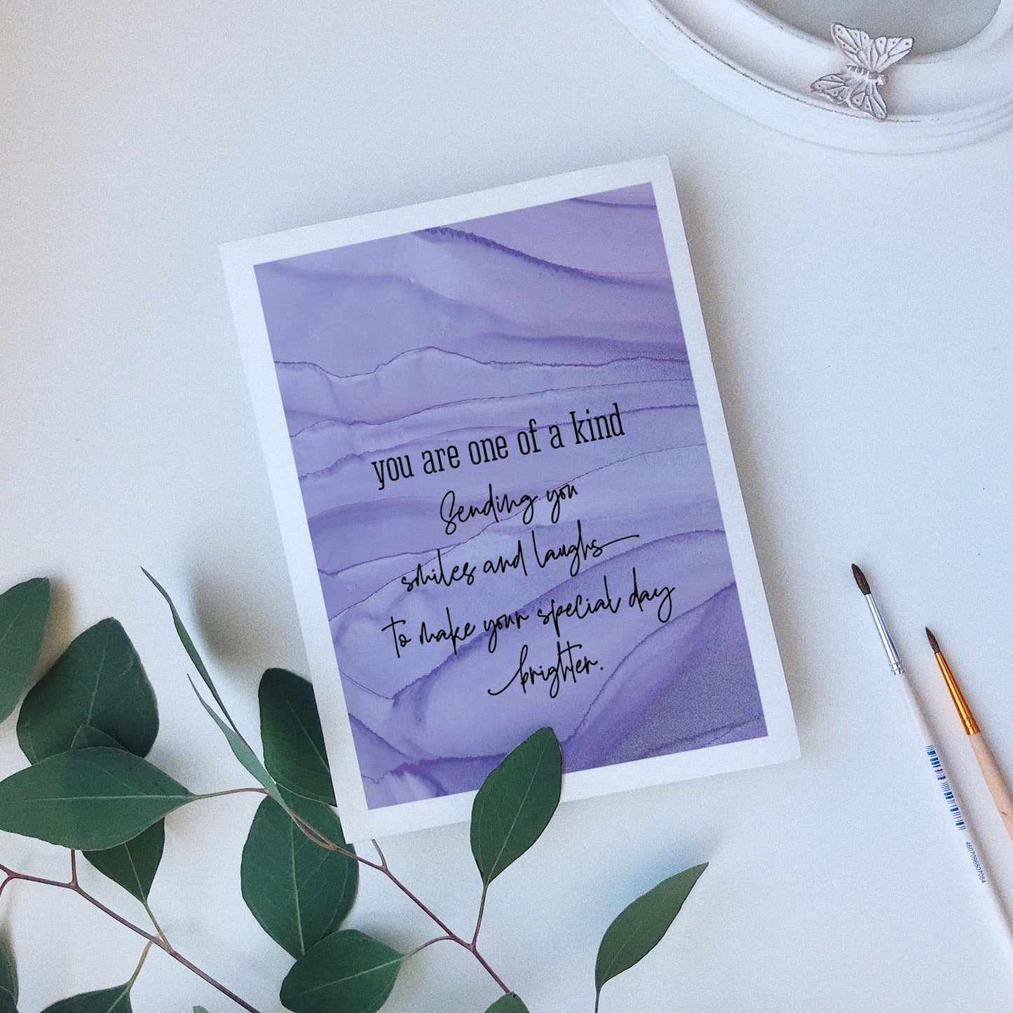 Greeting card laying on a white background near green leaves, paintbrushes and a ceramic dish.