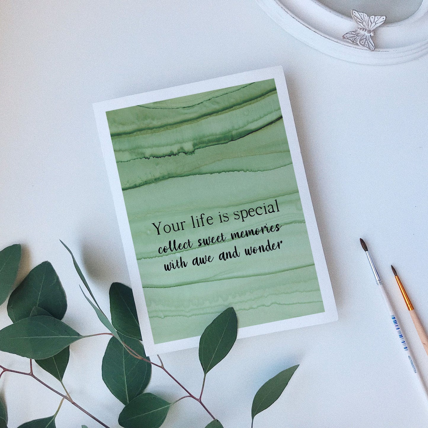 Greeting card laying on a white background near green leaves, paintbrushes and a ceramic dish.