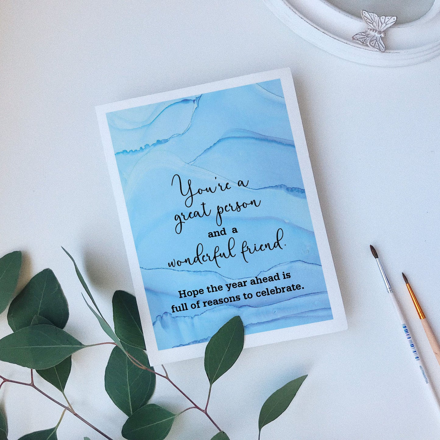 Greeting card laying on a white background near green leaves, paintbrushes and a ceramic dish.