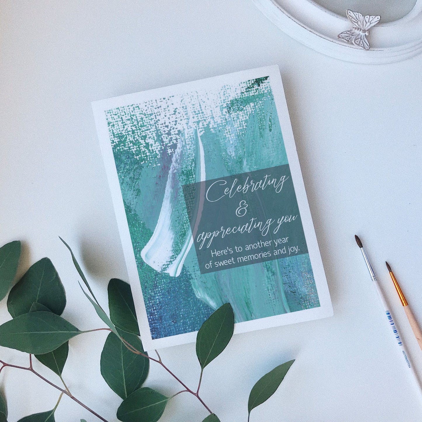 Greeting card laying on a white background near green leaves, paintbrushes and a ceramic dish.