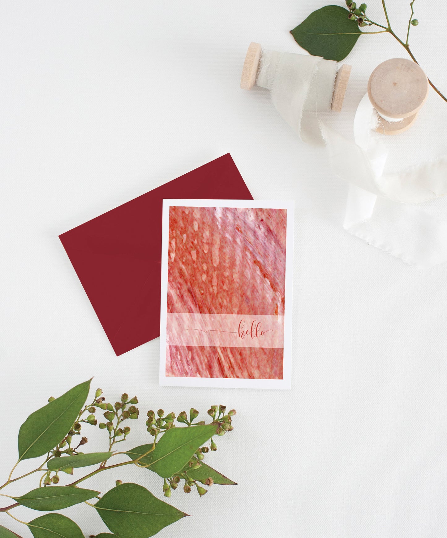 Greeting card with colorful envelope laying on a white surface with green leaves and cotton ribbon wound on wooden spools.