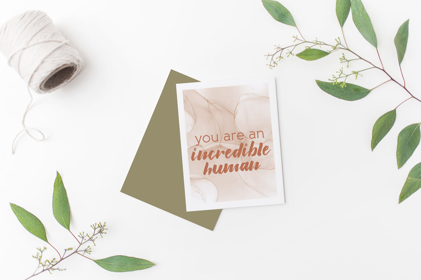 Greeting card laying on a white background near green leaves and a spool of white string.