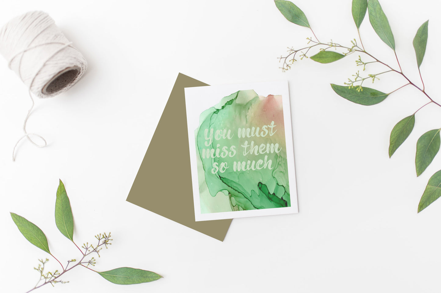 Greeting card laying on a white background near green leaves and a spool of white string.