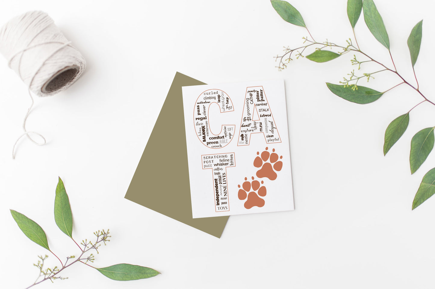Greeting card laying on a white background near green leaves and a spool of white string.