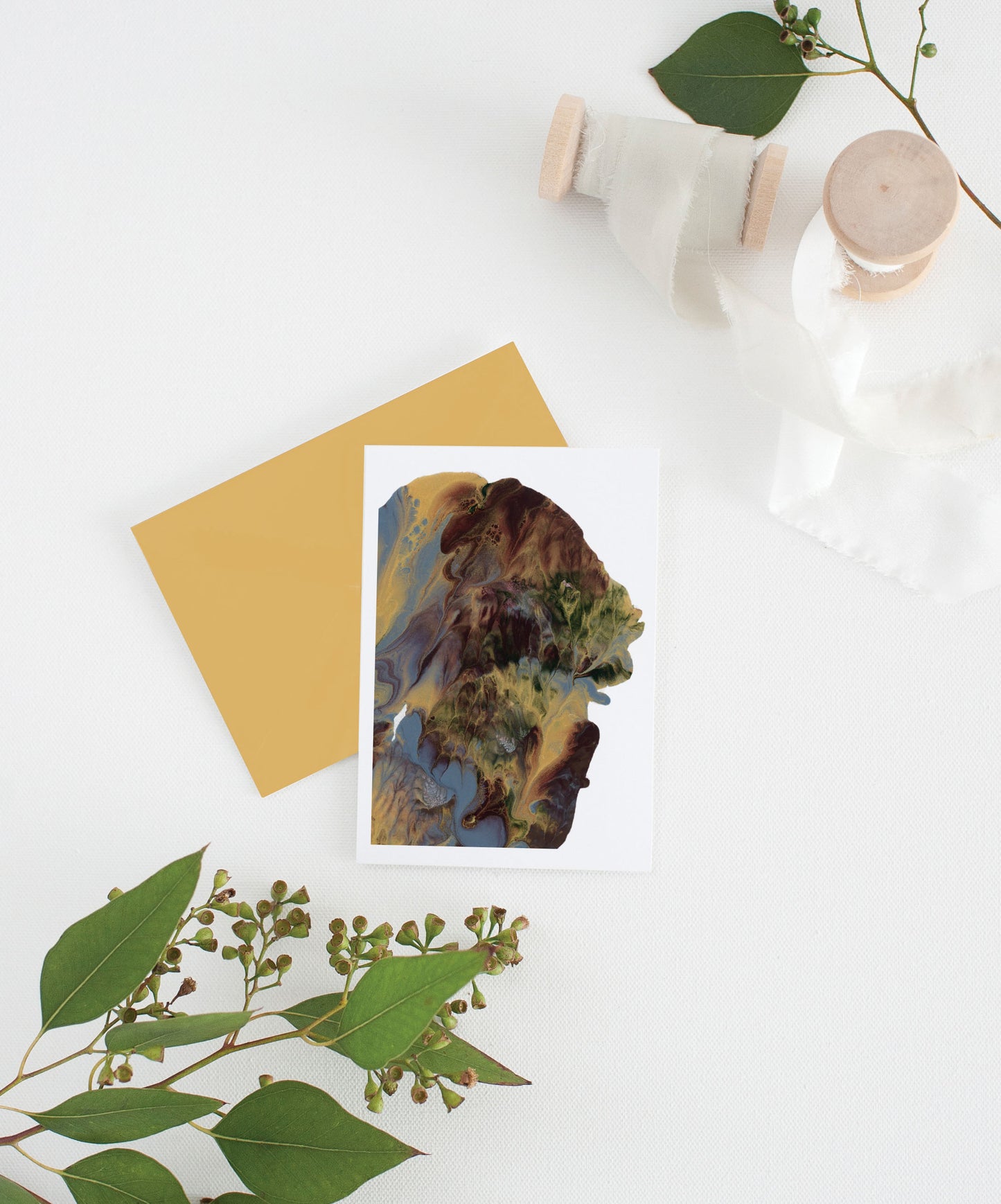 Greeting card with colorful envelope laying on a white surface with green leaves and cotton ribbon wound on wooden spools.