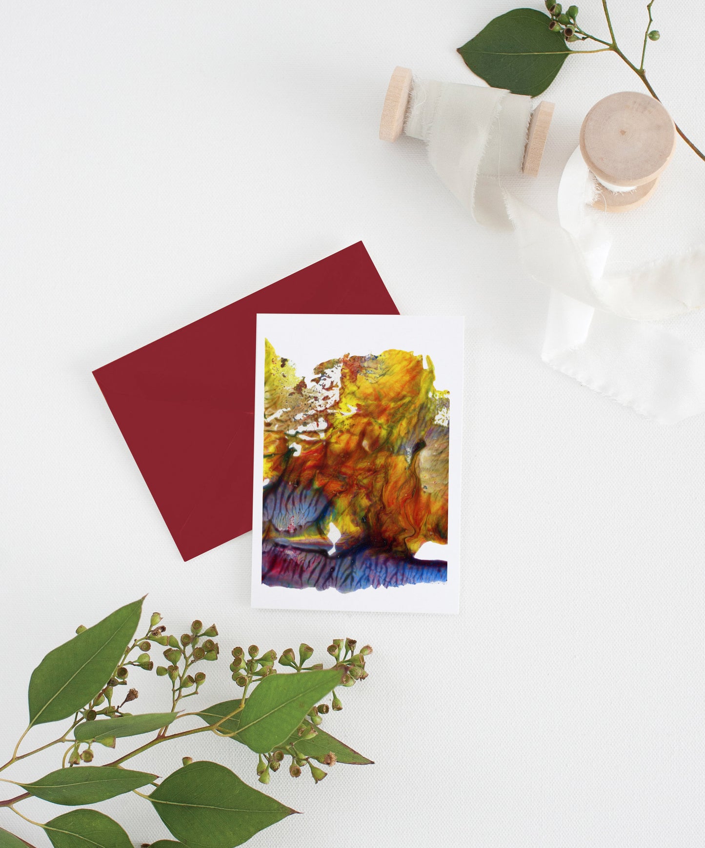 Greeting card with colorful envelope laying on a white surface with green leaves and cotton ribbon wound on wooden spools.