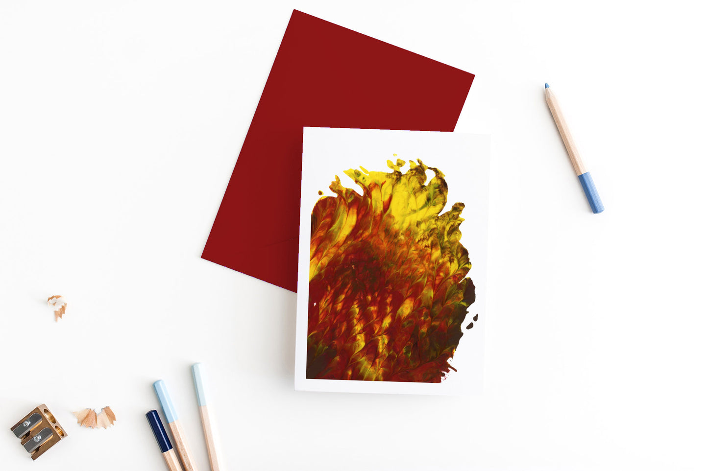 Greeting card showing an abstract painting in reds, yellows, and browns created from squishing acrylic paint between pieces of cardboard. It sits on a red envelope with 4 colored pencils, a sharpener and shavings on a white background.