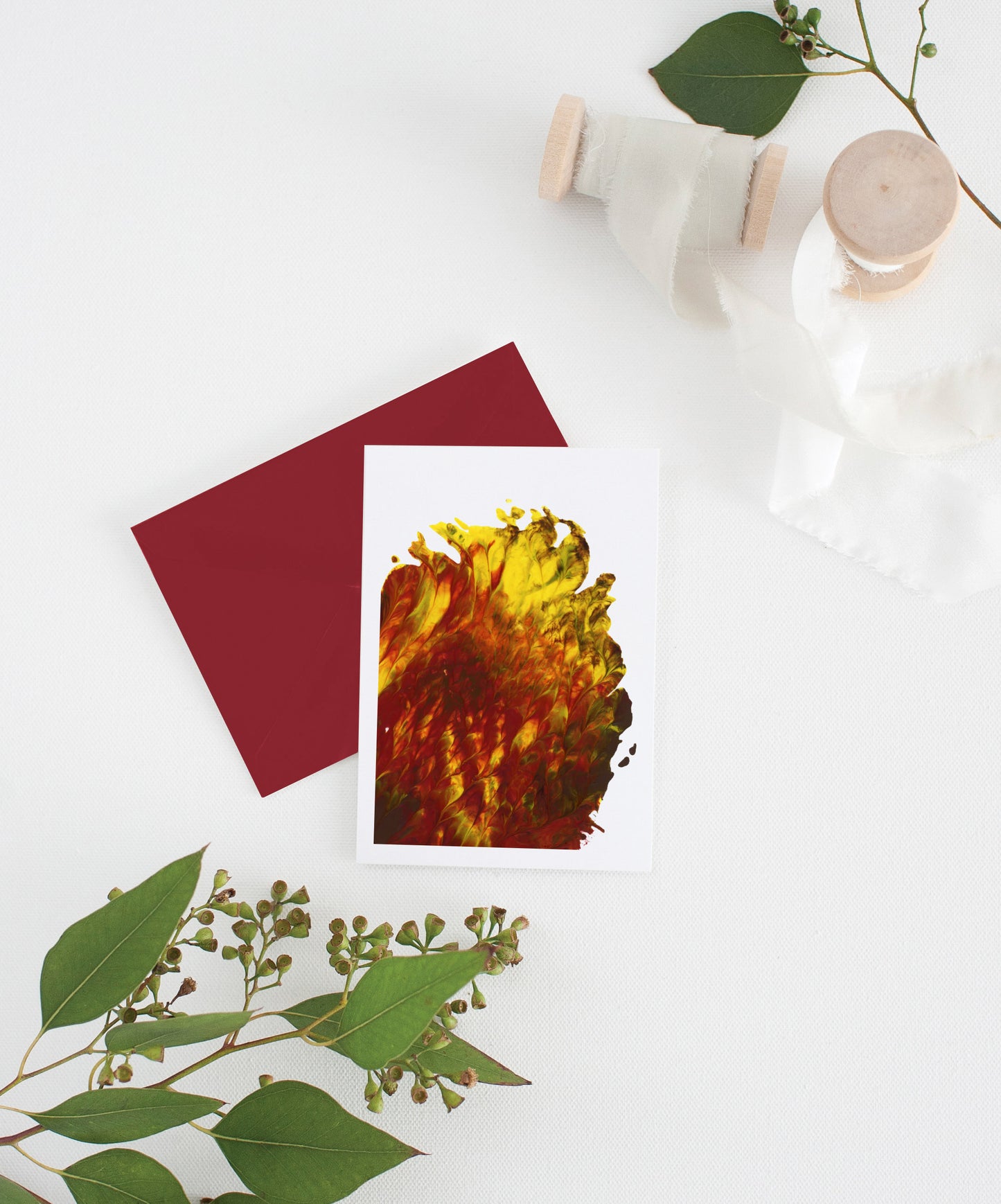 An abstract painting greeting card sits with a red envelope on a white background. There are leaves and two small spools of white fabric ribbon near it.