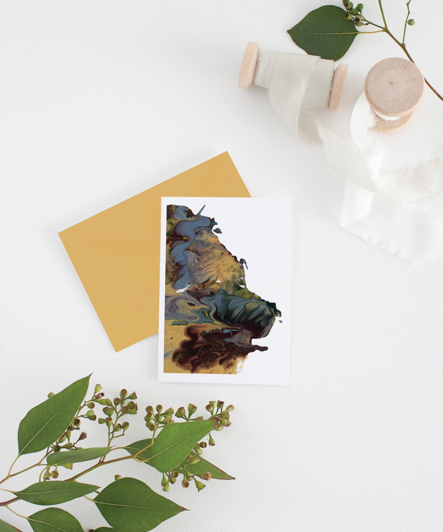 An abstract painting greeting card sits with a gold envelope on a white background. There are leaves and two small spools of white fabric ribbon near it.