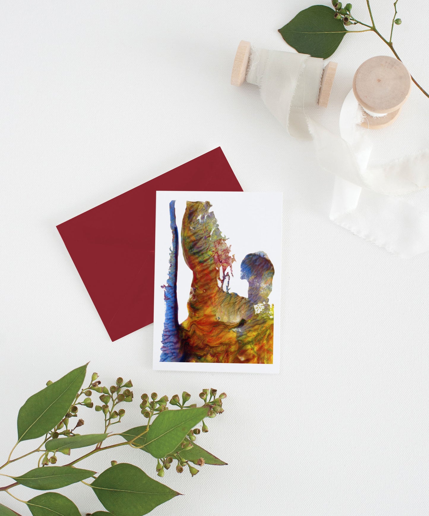 Greeting card with colorful envelope laying on a white surface with green leaves and cotton ribbon wound on wooden spools.