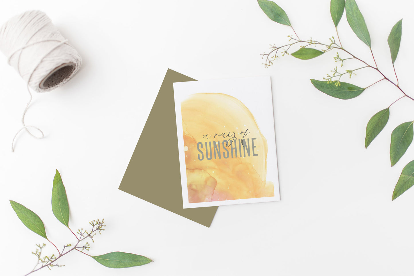 Greeting card laying on a white background near green leaves and a spool of white string.