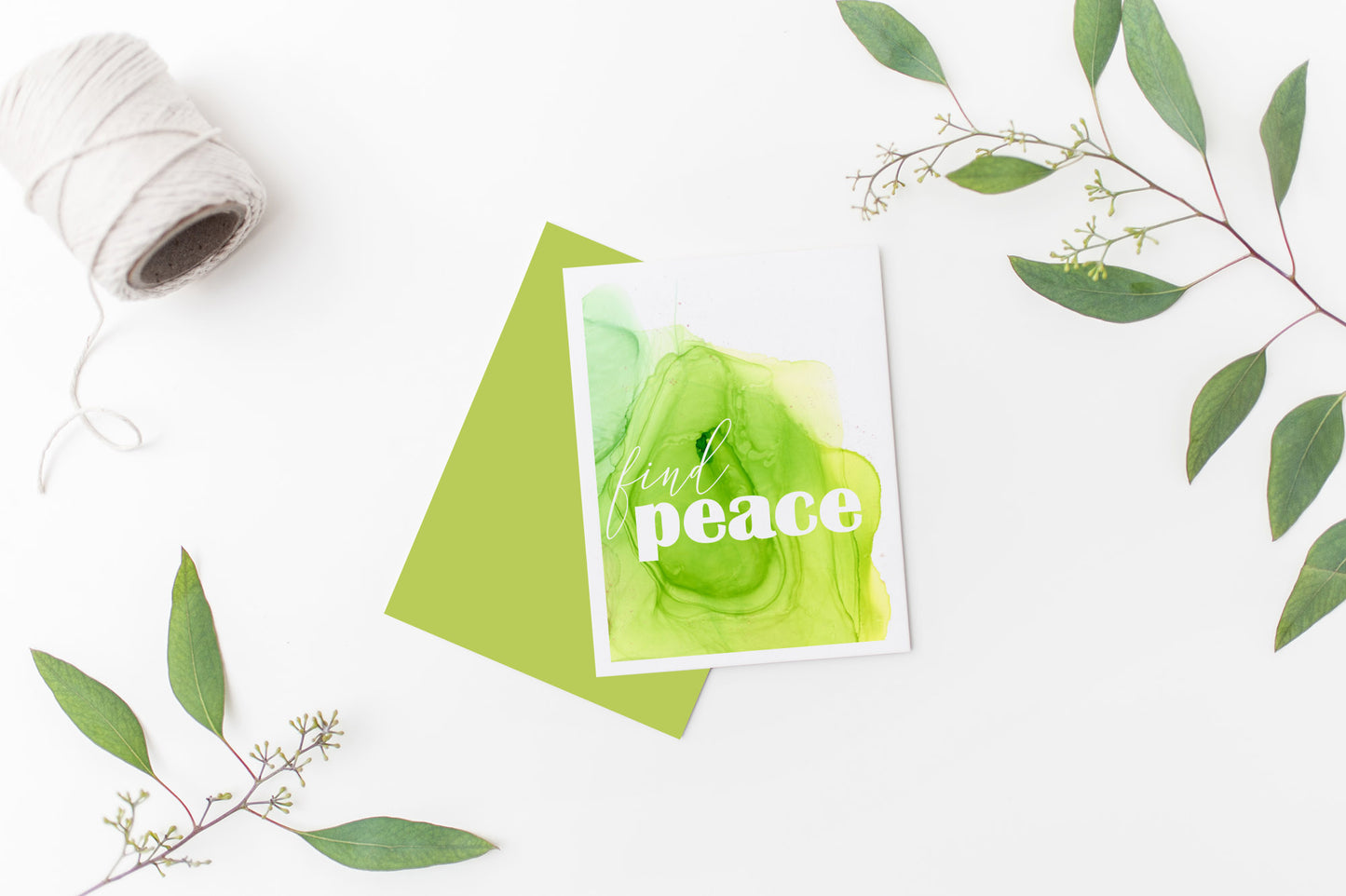 Greeting card laying on a white background near green leaves and a spool of white string.