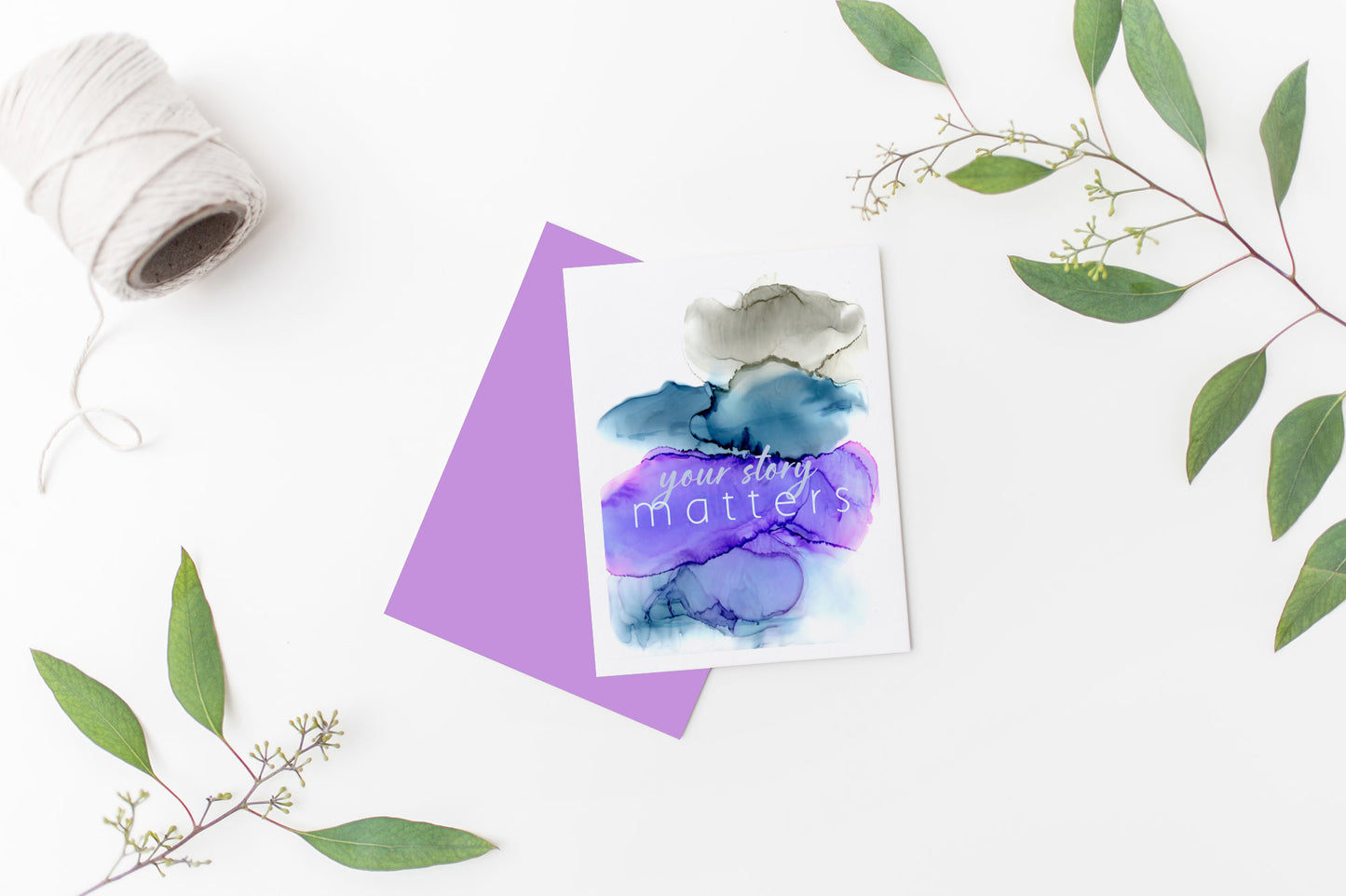 Greeting card laying on a white background near green leaves and a spool of white string.