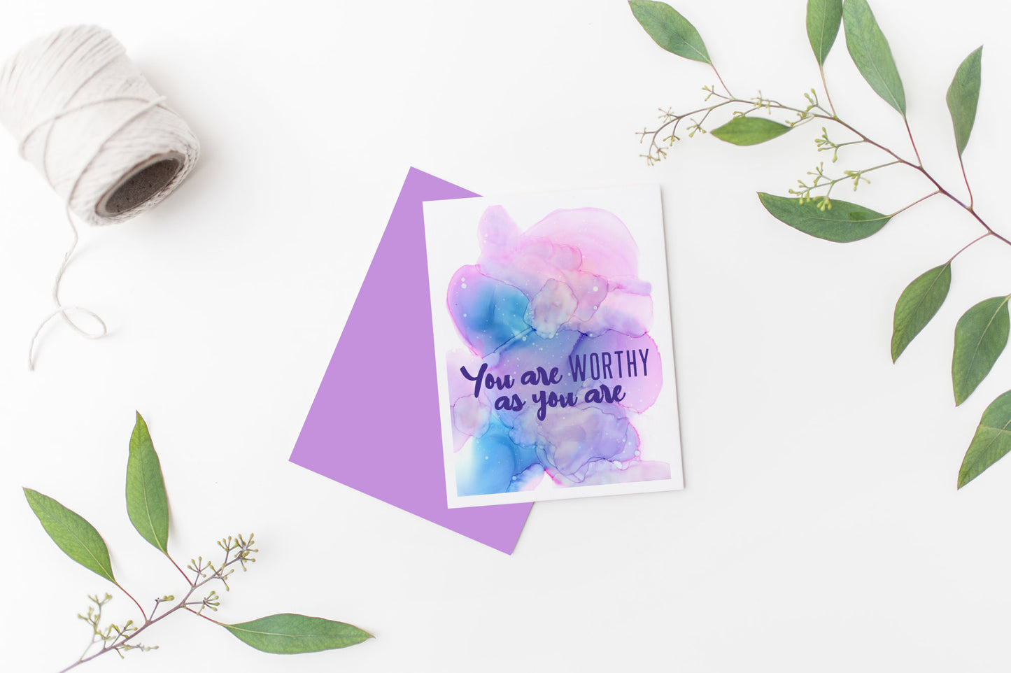 Greeting card laying on a white background near green leaves and a spool of white string.
