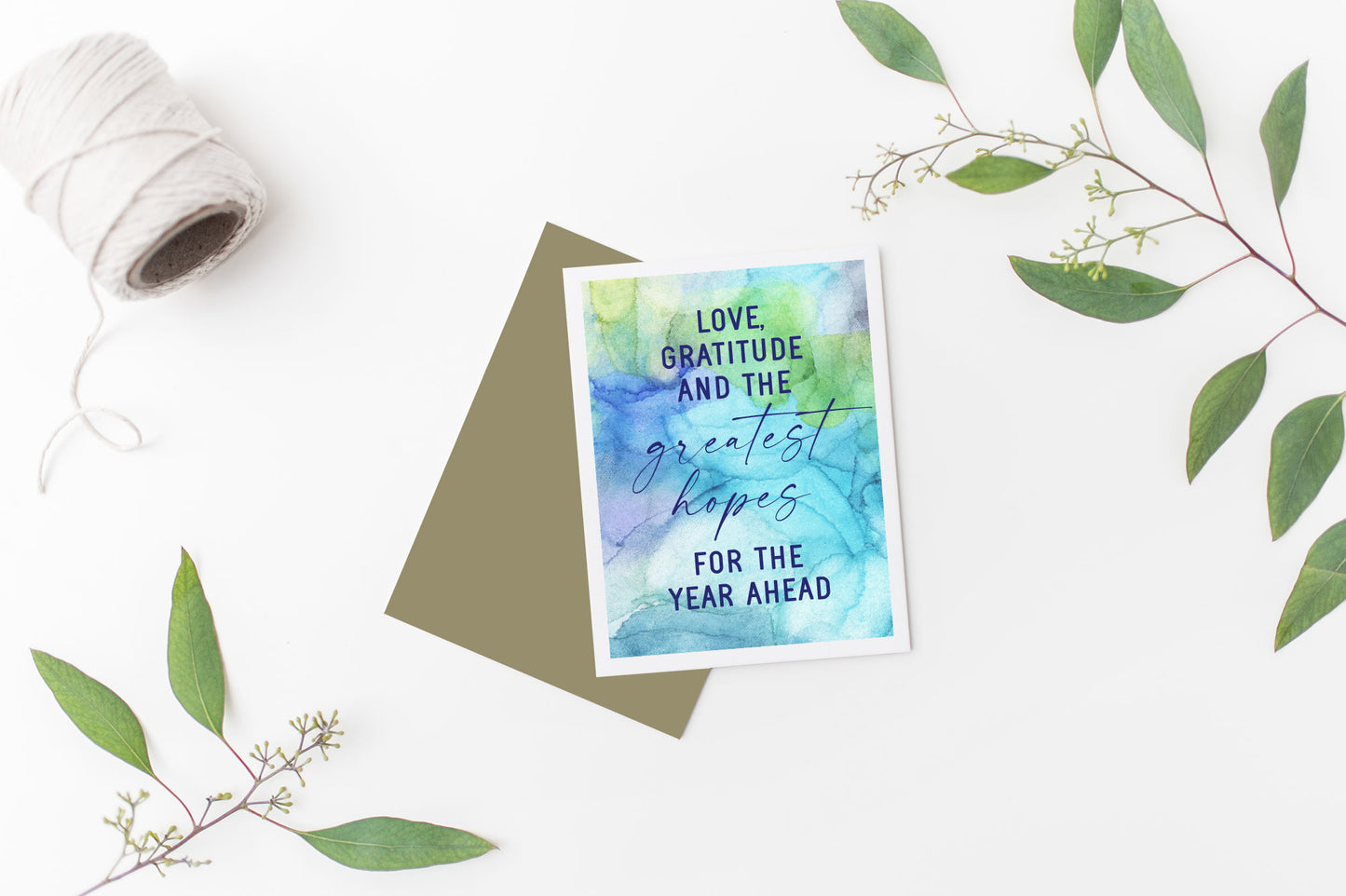 Greeting card laying on a white background near green leaves and a spool of white string.