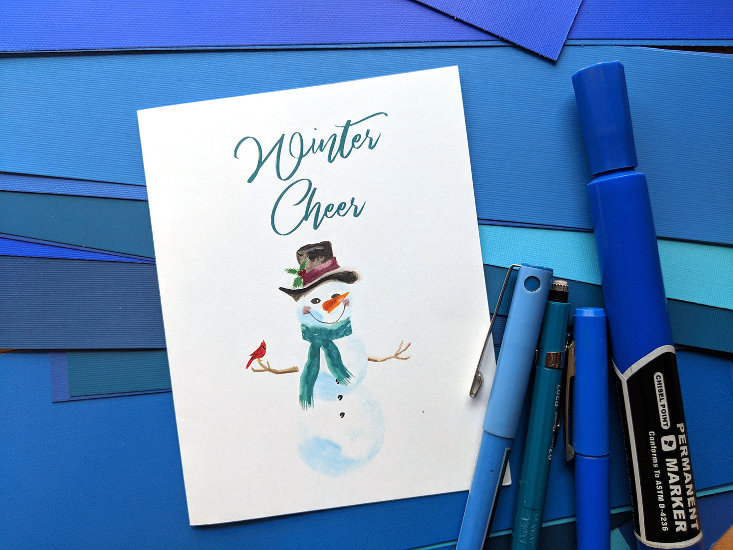 Greeting card on a fanned out stack of blue papers with four blue markers and pens.