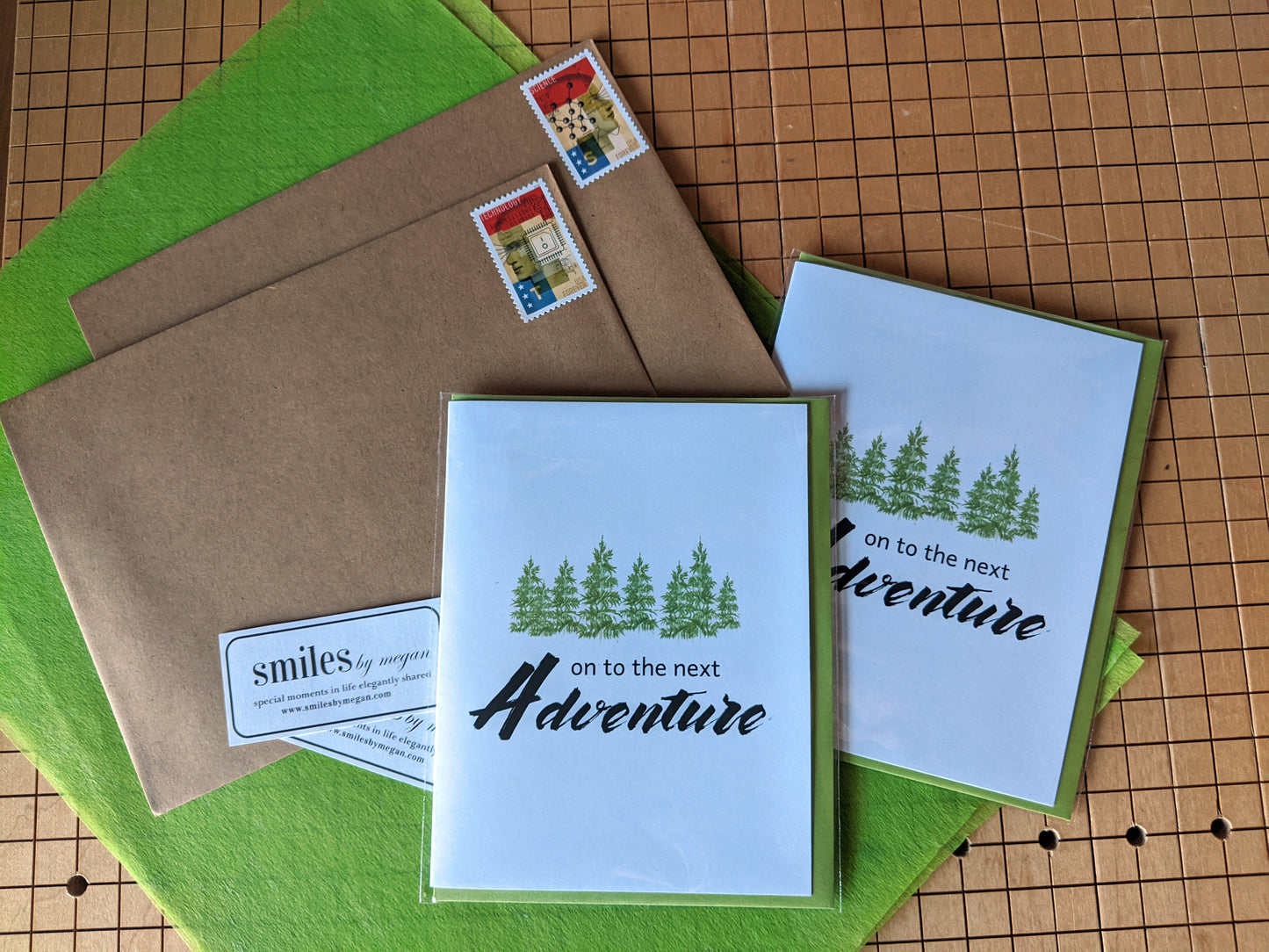 Greeting cards and kraft mailing envelopes, smiles by megan stickers, and green tissue laying on the grid of a paper cutter.