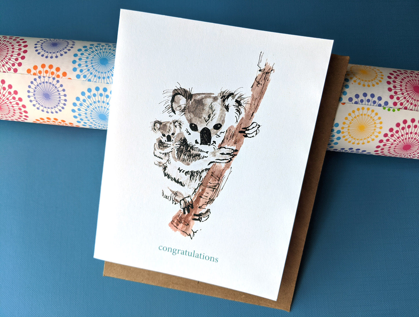 Greeting card with envelope leaning against a roll of colorful wrapping paper and a blue background.