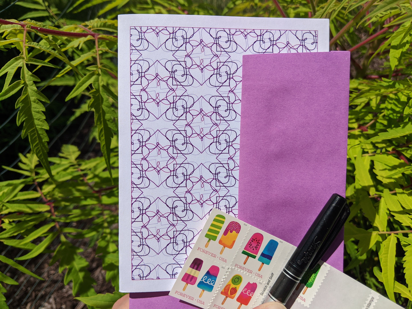 Hand holding a greeting card, colorful envelope, pen, and stamps in front of shrubs outside.