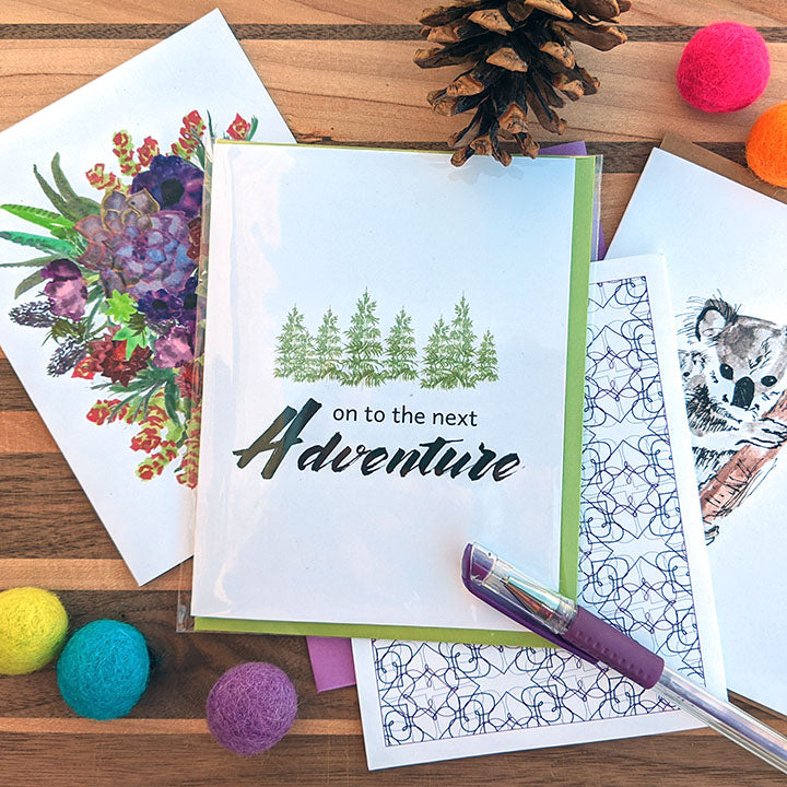 Overlapping greeting cards on a wooden surface with a pen, colorful felt balls, and pine cone.