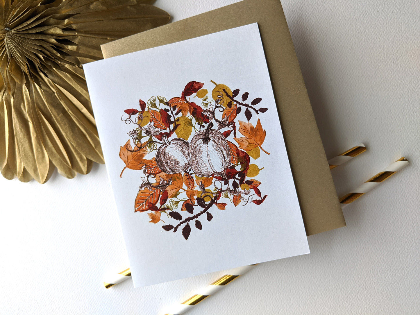 Greeting card and envelope leaning against a gold paper medallion and paper straws on a white background.