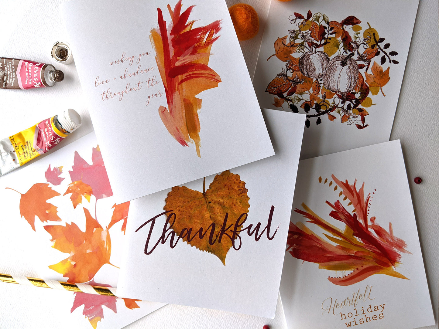 Five overlapping greeting cards on a white background with paint tubes, colorful felt balls, and a paper straw.