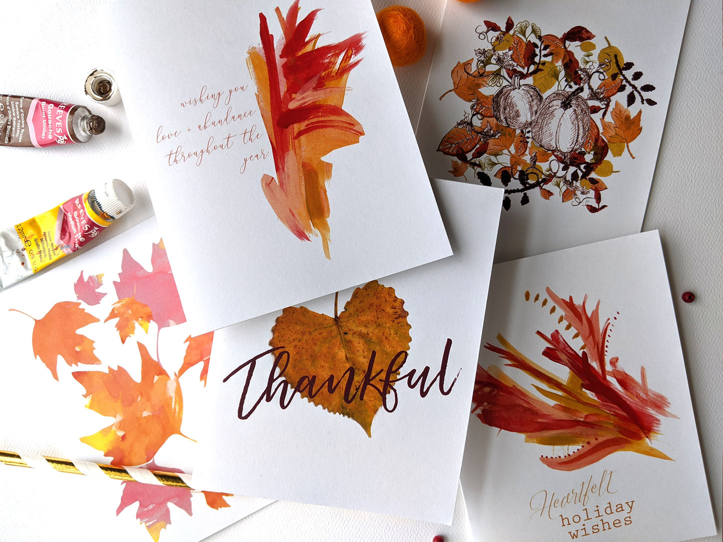Five overlapping greeting cards on a white background with paint tubes, colorful felt balls, and a paper straw.