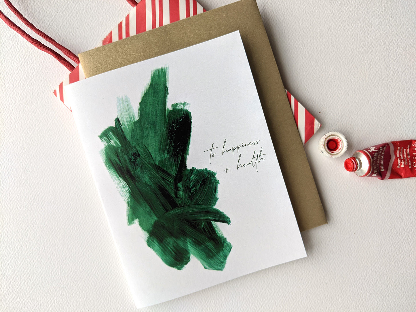 Greeting card with colorful envelope laying on a white surface with an open paint tube and striped gift bag.