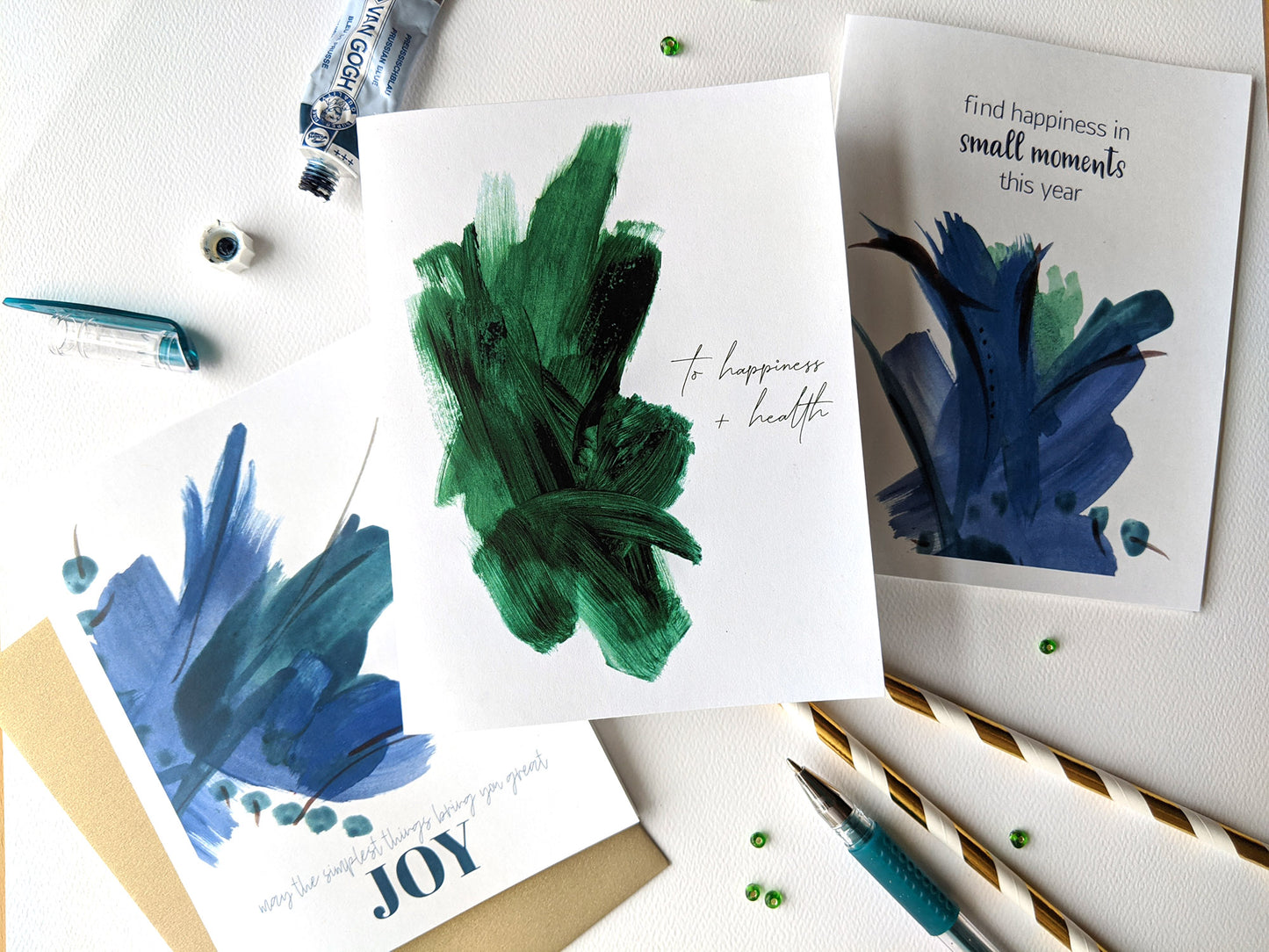 Three greeting cards overlapping on a white background with a tube of paint, pens, beads, and paper straws.