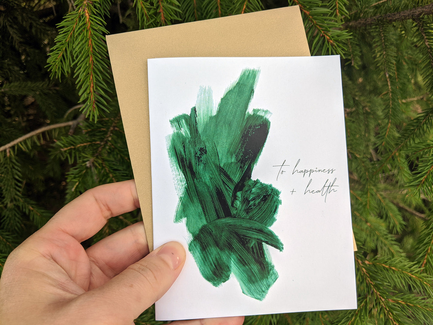 Hand holding a greeting card and colorful envelope in front of shrubs outdoors.