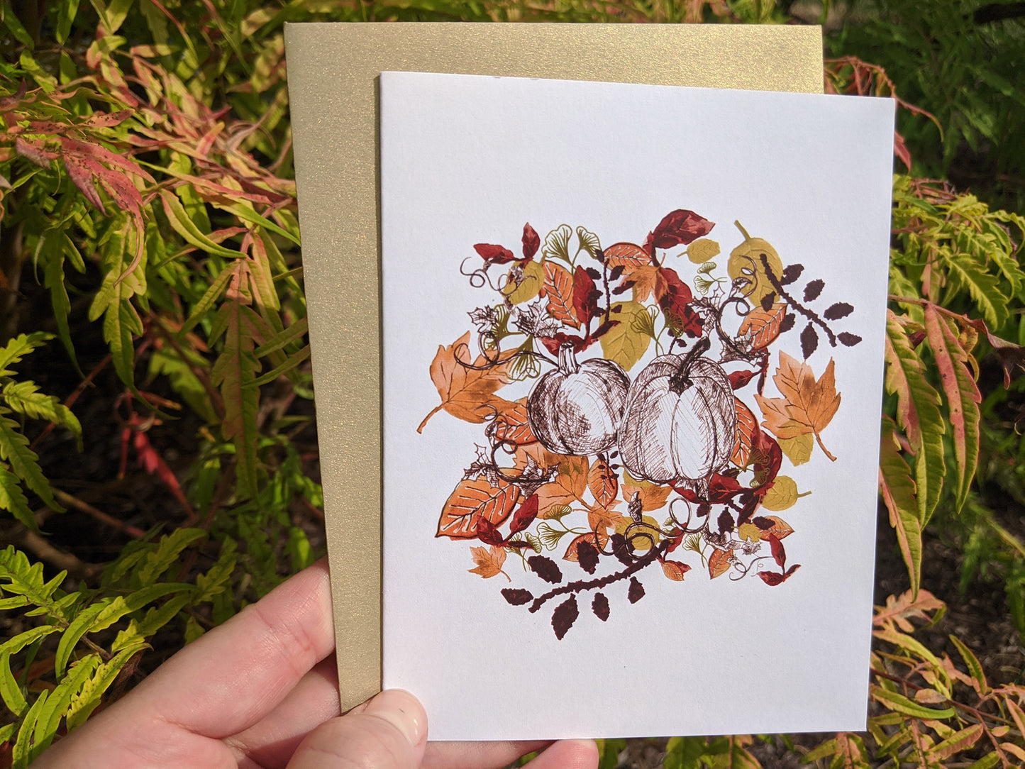 Hand holding a greeting card and colorful envelope in front of shrubs outdoors.